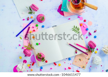 Colorful flat lay with an open book with blank pages, confetti, sweets, hard candy, pink envelope, macaroons, and tea. Feminine writer workplace with copy space.