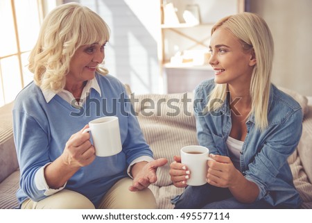 Beautiful old woman and young girl are drinking tea, talking and smiling while sitting on couch at home