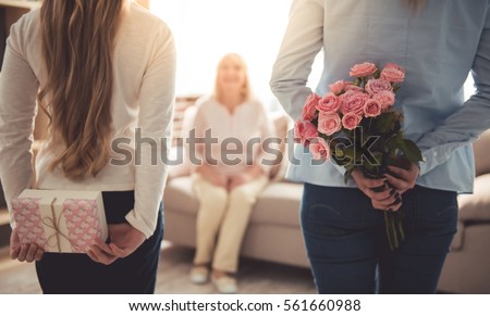 Teenage girl and her mom are hiding flowers and a gift box for their beautiful granny behind backs while grandma is sitting on couch at home