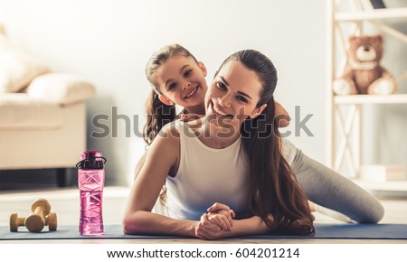 Beautiful young woman and charming little daughter are looking at camera and smiling while working out at home