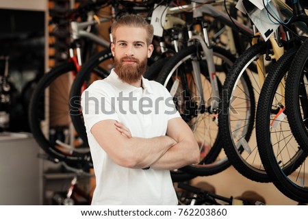 A salesman in a bicycle shop poses near a bicycle. There are parts, different types of bicycles. He smiles at the camera.