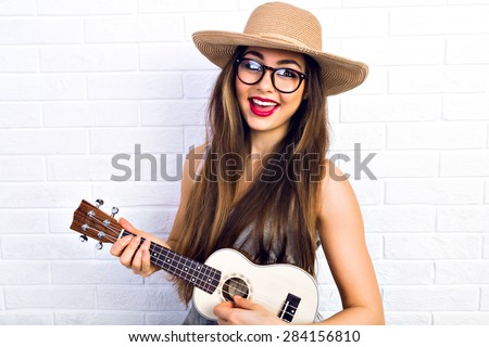 Young funny hipster girl having fun and playing on small ukulele guitar, singing and dancing. wearing vintage glasses and straw hat, joy, positive mood. White urban brick wall background.