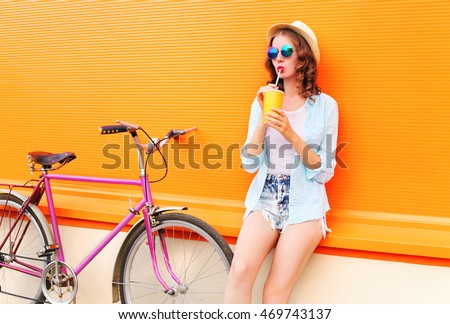 Fashion pretty woman drinks fruit juice from cup with retro bicycle over colorful orange background