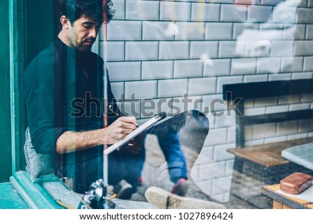 Pensive bearded young man dressed in black shirt writing down essay in notepad resting in coffee shop interior.Skilled hipster guy writer noting some records in notebook sitting in coworking space