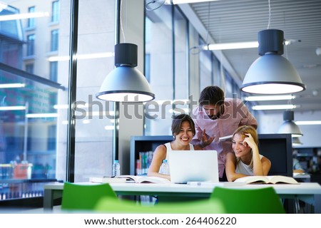 Group of international university students having fun studying in library, three colleagues of modern work co-working space talking and smiling while sitting at the desk table with laptop computer