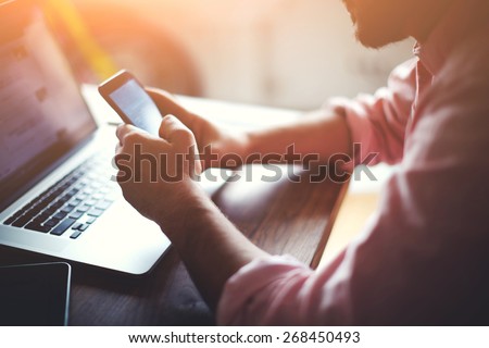 Silhouette of cropped shot of a young man working from home using smart phone and notebook computer, man's hands using smart phone in interior, man at his workplace using technology, flare light