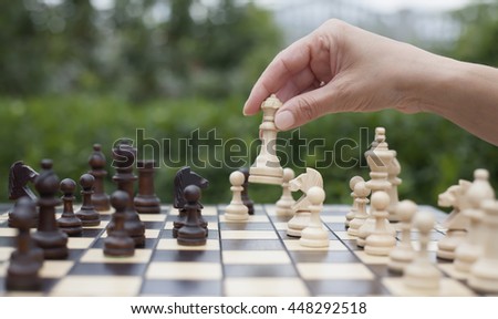 woman's hand moves a wooden piece of queen on the chessboard with other chess pieces on the green background of nature
