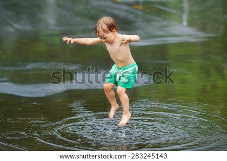 Portrait of happy toddler boy jumping in puddles during the rain thunderstorm on a bright summer day outside, sports recreation leisure concept, childhood and freedom