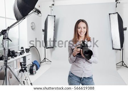 Young female photographer posing in the photo studio, she is smiling and holding a professional digital camera
