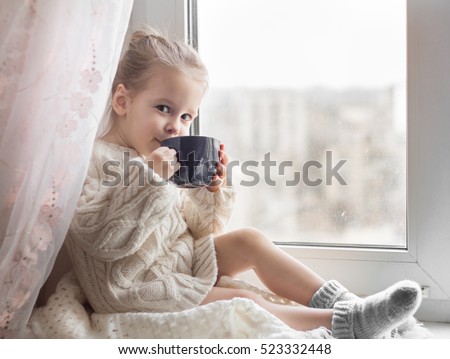 A little girl in the white knitted sweater sits on a window and drinks from a cup