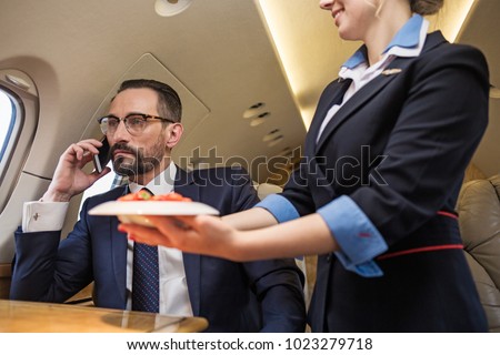Portrait of thoughtful good looking mature man talking by phone in airplane. Stewardess is serving plate of strawberries