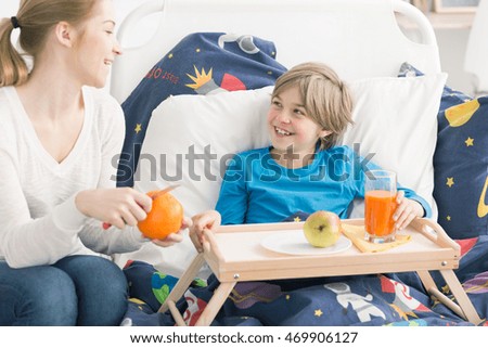 Smiling boy eating fruit and drinking juice in bed, on a bed tray, laughing with his mother