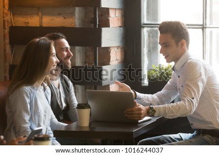Insurance broker or salesman making offer to young millennial couple using laptop in cafe, realtor consulting customers about mortgage sitting at coffeehouse table pointing on computer screen
