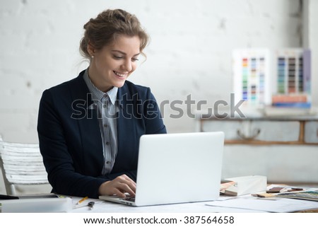 Portrait of beautiful happy smiling young designer woman working on laptop at office desk. Attractive cheerful model using computer. Typing indoors