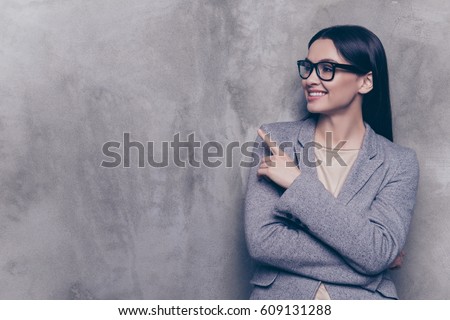 Portrait of smart young successful businesswoman in glasses and suit pointing to the side while standing near gray wall.