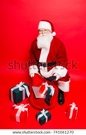 Holly jolly x mas noel! Busy santa in headwear, costume, black belt, shoes, white gloves fills sack with a lot of gifts for kids, with ribbon tapes bows, digital devices, electronics, playstation