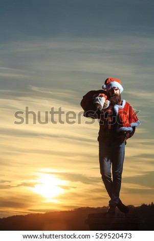 young handsome bearded bad santa claus man with long beard in checkered shirt jeans and red new year hat in christmas or xmas coat with toy sack on brick chimney in sunset, copy space