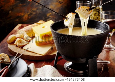 Gourmet Swiss fondue dinner on a winter evening with assorted cheeses on a board alongside a heated pot of cheese fondue with two forks dipping bread and white wine behind in a tavern or restaurant