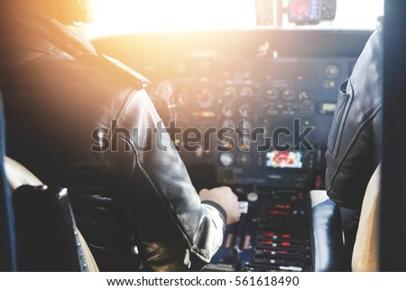 Two unrecognizable jet airliner pilots wearing leather jackets piloting aircraft at sunset, sitting inside cabin at steering control with modern dashboard, sun shining through cockpit windscreen