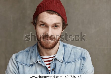 Close up shot of charismatic fashionable young European male with thick beard wearing trendy hat and denim jacket, staring at camera and smiling with happy cheerful look, enjoying free time at cafe