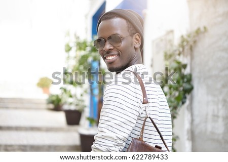 People, lifestyle, leisure, tourism and travel. Outdoor shot of dark-skinned male student smiling broadly, enjoying good sunny day walking on streets of European city, wearing stylish clothes
