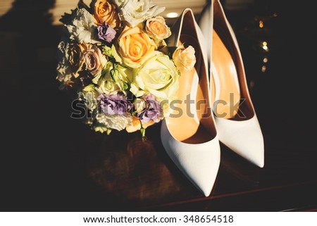 Luxury white leather women's high-heeled shoes on the table next to the magnificent colorful wedding bouquet of beautiful roses.