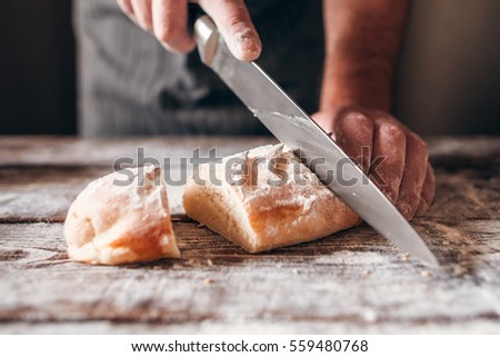 Cutting fresh crusty bread close-up. Baker carving loaf of warm soft tack with big knife on kitchen table. Dinner preparing, homemade bakery, traditional recipe concept