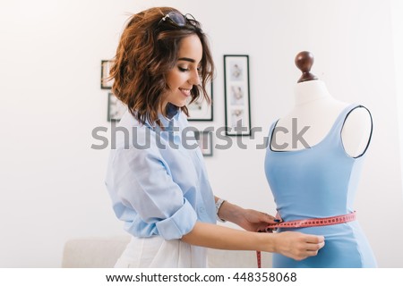 A brunette girl in a blue shirt is working in the workshop studio. She makes fitting on the blue dress on the mannequin.