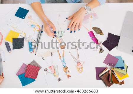This picture describes the processes of designing clothes. There are hands of a girl drawing sketches on the table. There is creative mess with different stuff on the table.