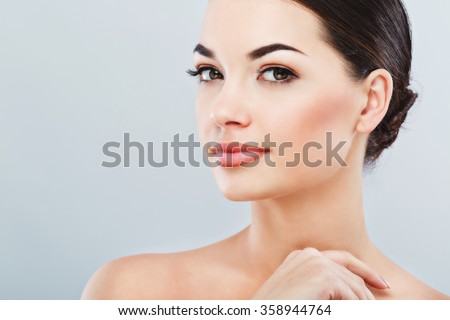 Pretty girl with big eyes and dark eyebrows, with naked shoulders, looking at camera and smiling, a model with light nude make-up, gray studio background, beauty photo, copy space, close up.