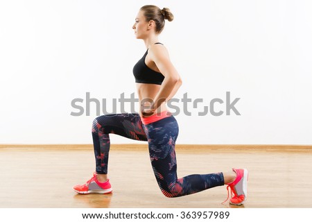 Sport girl with dark hair wearing pink snickers, dark leggings and black short top doing lunge at gym, fitness, white wall and wooden floor, copy space.