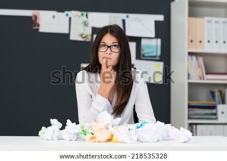 Young woman with writers block sitting in an office with a desk littered with crumpled paper as she sits looking thoughtfully into the air with her finger to her chin seeking new ideas