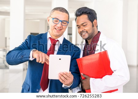 Young Indian doctor with businessman talking in hospital hallway - Salesman holding pad showing health care equipment at asian medical director - Concept of sales, business, deals with multiracial men