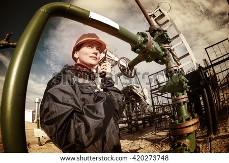 Female worker in the oil field talking on the radio wearing orange helmet and blue work clothes. Industrial site background. Toned.