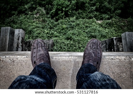 Suicide concept., Depressed young man looking down at his shoe and contemplating suicide., On the edge of a high bridge with forest below.