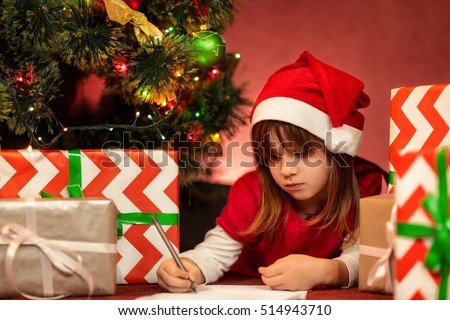 Christmas routine. Portrait of a serious little girl in a red dress and Santa Claus hat lying on the floor under the Christmas tree writing a letter