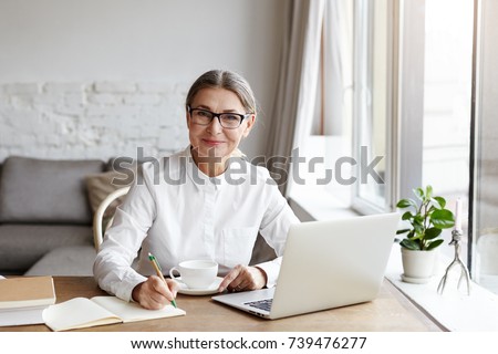 Attractive positive talented mature woman writer sitting in front of laptop and writing her new book about love, looking at camera with joyfull smile. Senior female teacher making notes in copybook