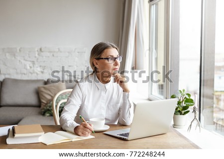 Attractive blonde mature female writer sitting at desk at home placing chin on her hands and looking away with thoughtful or unhappy expression while experiencing writer's block and creative slowdown