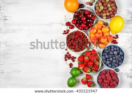 Various fresh fruits in bowls on white wooden background with copy space: strawberries, blueberries, raspberries, cherries, red currants, gooseberries, apricots, orange, lemon and limes. Top view.