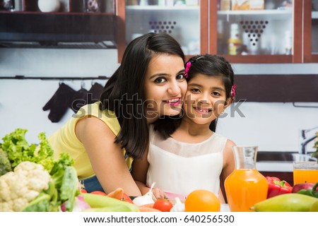 indian mother and daughter in kitchen, with table full of fruits and vegetables. both are busy either in chopping, eating, drinking, mixing something