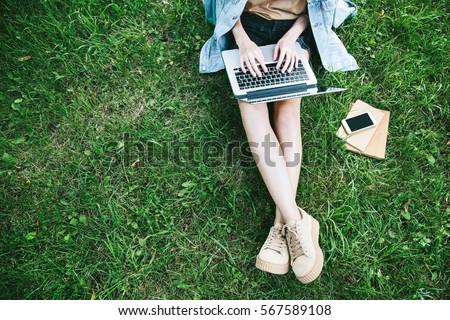Top view of woman sitting in park on the green grass with laptop, notebook and smartphone, hands on keyboard. Computer screen mockup. Student studying outdoors. Copy space for text