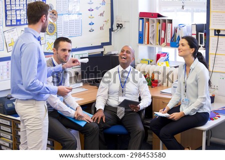School teachers gather in a small school office for a chat. They look happy. A woman and three men group together.