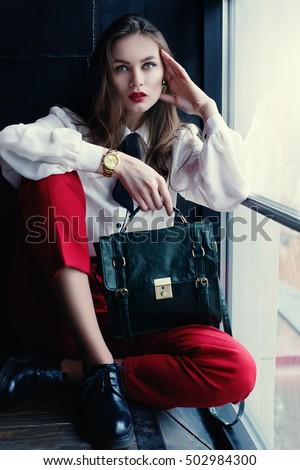 Indoor full body portrait of young fashionable woman posing in loft interior. Model looking at camera. Lady wearing stylish clothes and holding green handbag. Female fashion concept. City lifestyle.
