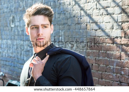 Handsome young man walking in an urban street with his suit jacket slung over his shoulder as he looks away, close up upper body view. Attractive male hipster model.