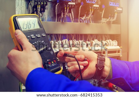 Multimeter in hands of electrician close-up against background of electrical wires and relays. Adjustment of scheme of automation and control of electrical equipment