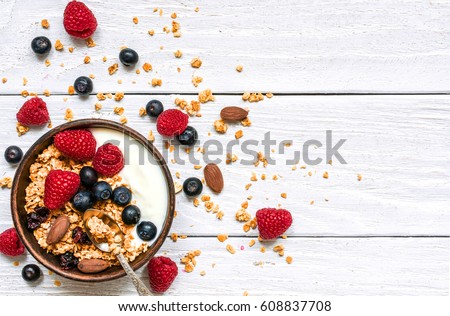 bowl of oat granola with yogurt, fresh raspberries, blueberries and nuts with spoon on white wooden board for healthy breakfast, top view