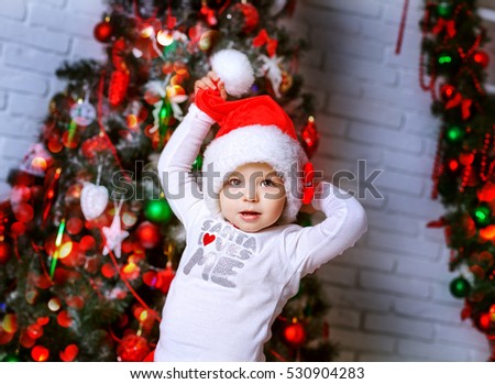 A little adorable girl in red santa's suit in a christmas decoration having fun . Cute children with toys indoors near xmas tree smiling. New year photo.