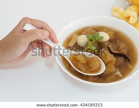 An Asian-forties-lady's hand is using a spoon to scoop Chinese style soup; or braised fish maw in red gravy with eggs, mushroom, and bamboo shoot in white bowl. White background.