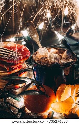 A mug with cocoa and melted marshmallows with a garland, burning Bengal lights and sparks, spruce branches, mandarins and pancakes with jam on a wooden table. Hot chocolate. Christmas miracle