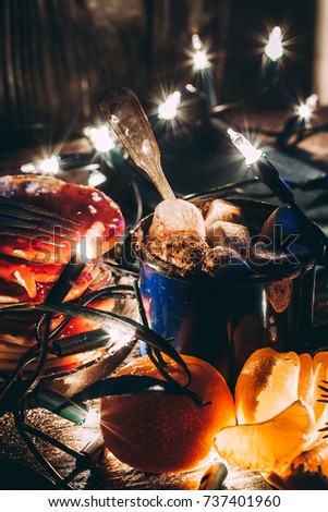 Mug with cocoa and melted marshmallows with a light Christmas tree garland, spruce branches, mandarins and pancakes with jam on a wooden table. Hot chocolate. Christmas cosiness.